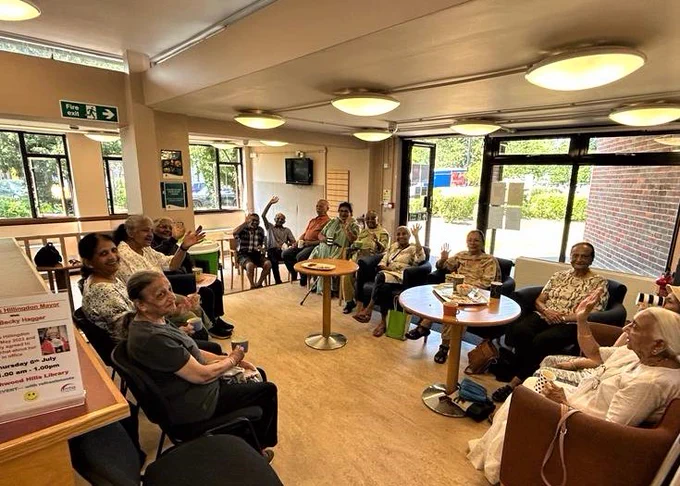 Photograph taken within Northwood Hills Library of their Gujarati Meet Up group. The camera is positioned at a standing level looking down on the group who are seated in chairs arranged in a rectangular shape. The attendees are looking up towards the camera, waving and smiling. In the middle of the group there are two light wooden coloured round tables.