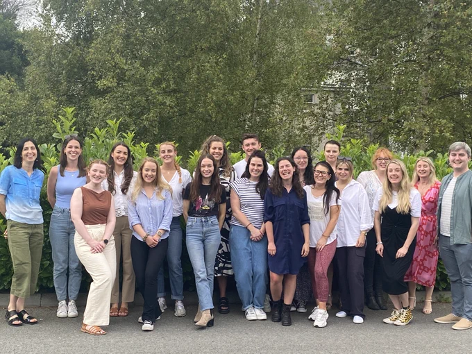 Class photo of students and staff from UCD. Group of approx 20 people are standing outside next to greenery. 