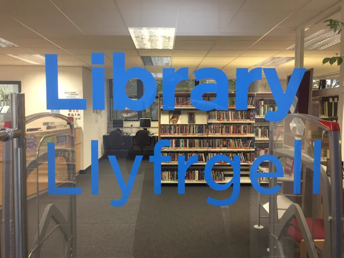 An image of the view through the library door. Library / Llyfrgell is written on the glass.