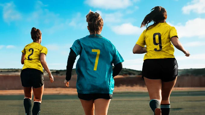 Three women in football strips are running towards an open field.
