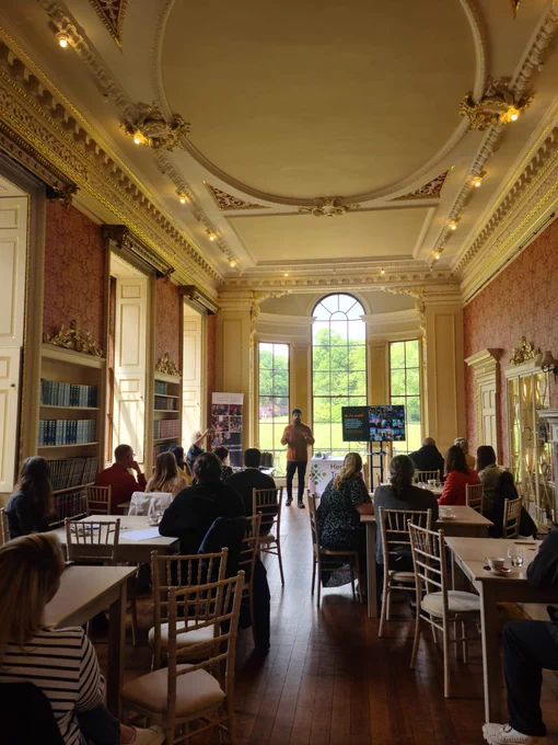Image is from Heritage Trust Network event with Rebel Creatives at Wentworth Woodhouse. Amrit is standing infront of a bay window in a room adorned in period coving. In the foreground, participants of his workshop are sitting on tables with their backs to the camera, engrosed in Amrits workshop.