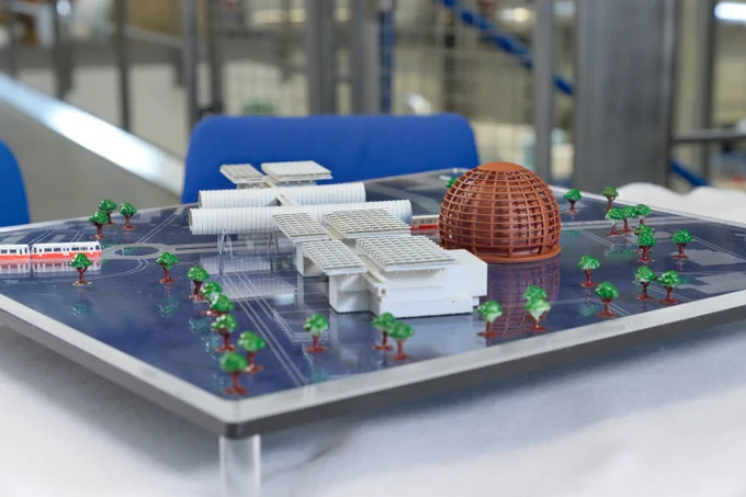 Image of a plastic mock-up model of CERN Science Gateway, almost LEGO-like. Five white buildings, two of which are shaped like horizontal tubes, are interconnected by bridges. Next to them, on the right side, there is a miniature version of a sphere-shaped building made out of timber, the Globe of Science and Innovation. 