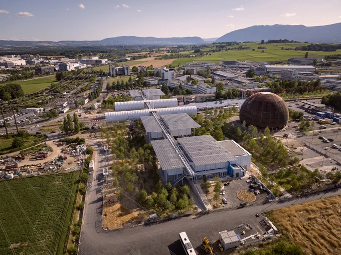 Image of five white buildings surrounded by trees. Two buildings are shaped like horizontal tubes, interconnected by a bridge that crosses over a traffic road. Next to them, on the right side, there is a sphere-shaped building made out of timber, the Globe of Science and Innovation. 