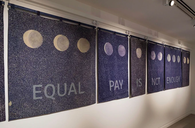 Six mottled blue banners hanging in a row on a white wall. The top of the banners have part of the cycle of the moon over 13 moons. At the bottom of the banners  there is some text that reads "EQUAL PAY IS NOT ENOUGH"