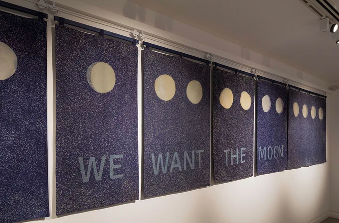 Six mottled blue banners hanging in a row on a white wall. The top of the banners have part of the cycle of the moon over 13 moons. At the bottom of the banners  there is some text that reads "WE WANT THE MOON"