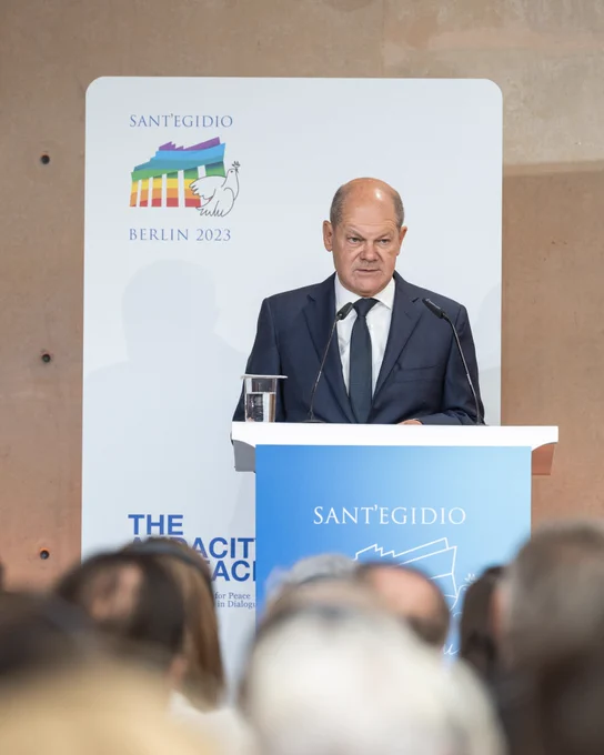 Bundeskanzler Scholz bei seiner Rede beim 37. Internationalen Friedenstreffen der katholischen Gemeinschaft Sant’Egidio in Berlin, am Rednerpult stehend. Im Hintergrund ist das Logo der Veranstaltung zu sehen.