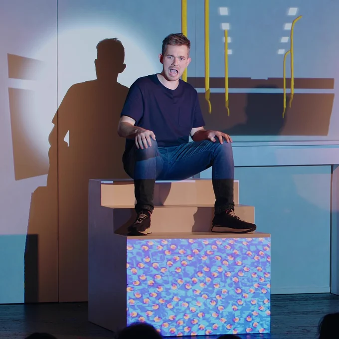 image shows someone wearing navy top and jeans sitting on a box on stage looking shocked