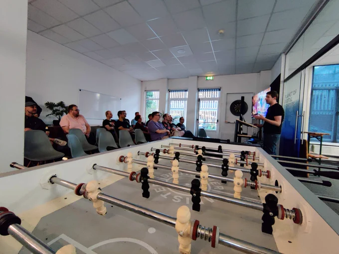 Mensen luisteren naar een presentatie van Robert Slootjes. Tafelvoetbaltafel staat op de voorgrond.