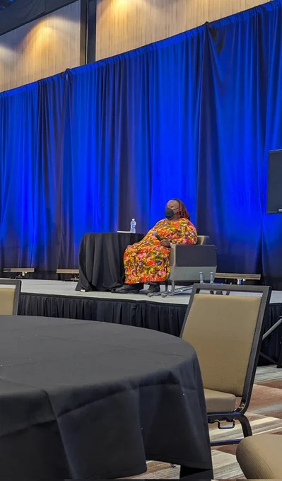 Black woman seated on a stage. She is wearing a mask and a pink and red floral gown. 