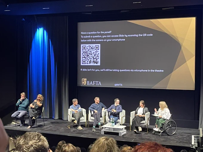 Five people sit for a Q & A panel on the stage at BAFTA. There is a blue curtain in the background and a screen with words in INTT audience members to ask questions. Two people also sit to the left.