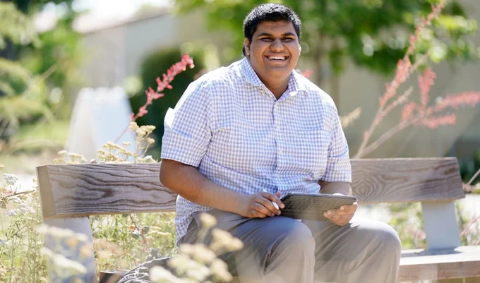 A photo of Ali Dadawalla sitting on a bench on Fresno State campus. He is smiling and it is bright and sunny.