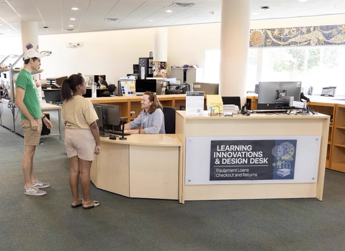 A tall man and a shorter woman stand in front of a smiling staff member at the Learning Innovations and Design Desk.