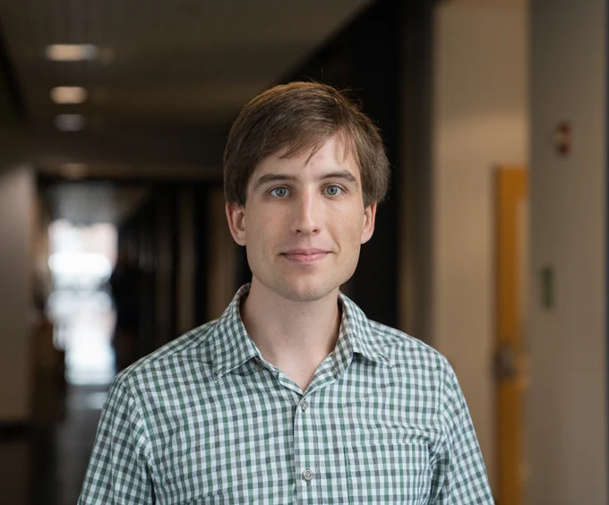 Male accelerated graduate student at UNH wearing a green checkered shirt in a hallway with a blurred background.