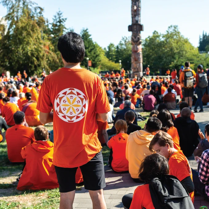 A crowd of people wearing orange shirts gather in front of The Reconciliation Pole at UBC.