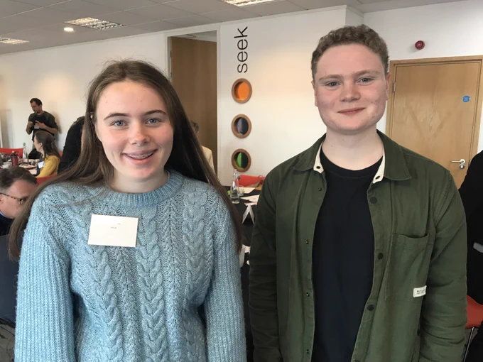 Two young people stand together. There is is teenage girl with long brunette hair and a blue jumper on the left, and a young man with short hair and a khaki jacket on the right.