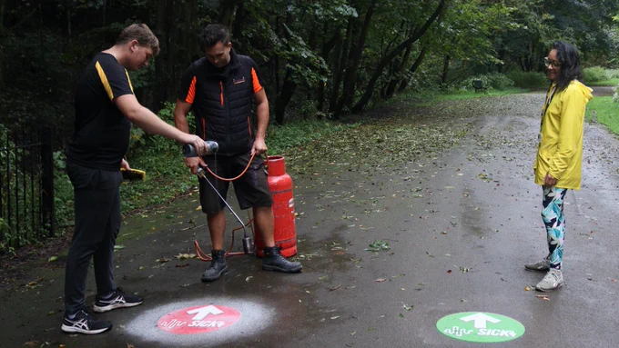 Image two shows two males from funactiveplaygrounds installing a red Soundpath marker next to a pre-exisiting green marker from the other route. Danielle is watching this task be completed and making sure the signs were pointing the correct way. It was a very wet day so the sky is grey and the concrete floor is dark due to the rain.