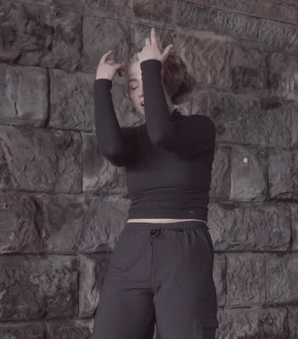 A performer wearing all black lifts their arms in front of their face in front of a stone wall.