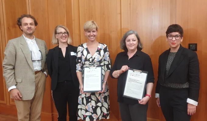 Vier Personen sind auf dem Bild, links ein Mann und drei Frauen, alle in förmlicher Kleidung. Zwei Frauen in der Mitte halten den offenen Brief der digitalen Zivilgesellschaft über den Global Digital Compact in die Kamera