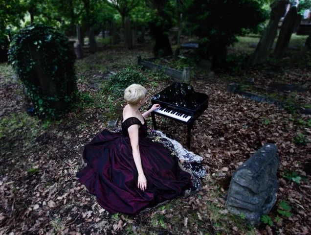 A gothic picture of Kate in a flowing velvety purple dress with one of her toy pianos in a graveyard