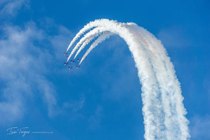Red Arrows in formation with a trailing arch of white smoke against a blue sky
