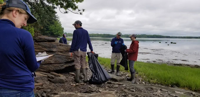 A group of people removing trash from a rocky coastline with trees and water in the background.