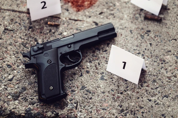 A black handgun lays on the ground next to an evidence marker with the number one on it. There is also a bullet and cartridge casing by other evidence markers and a red smudge on the ground near the top of the photo.