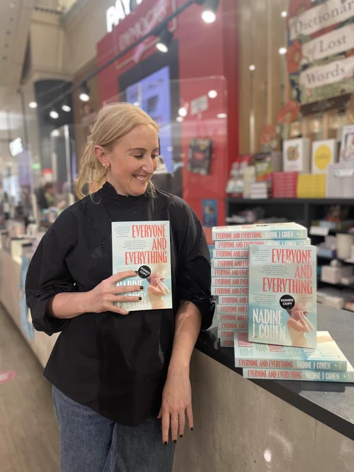 Author and former Twitter aficionado Nadine J. Cohen holding up her debut book, Everyone & Everything. There is a glorious blue stack of books next to her, with stickers that say "Signed by the author"