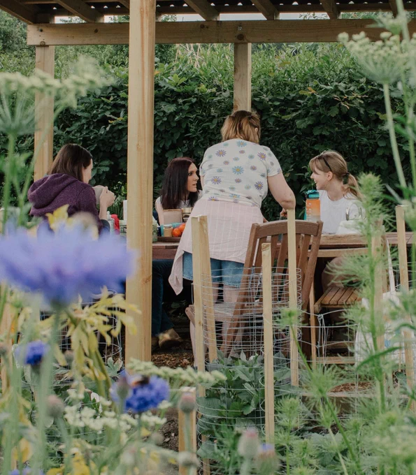 A group of 4 women are sitting under a wooden structure chatting and laughing together. On the table is a kettle, some teabags, some fruit and a water bottle. Out of focus in the foreground of the image, there are some beautiful blue wildflowers. 