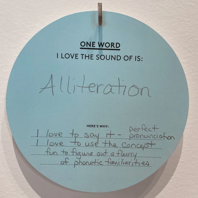 A circular blue card hanging on a peg on a wall. Text on the card reads: One word I love the sound of is: “Alliteration” Here’s Why: “I love to say it — perfect pronunciation, I love to use the concept, fun to figure out a flurry of phonetic familiarities”