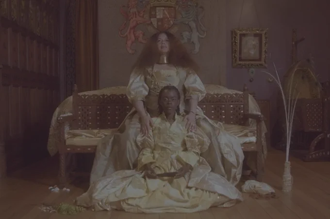 A film still shows two women dressed in golden dresses sat in an ornate room and looking boldly at the camera. 