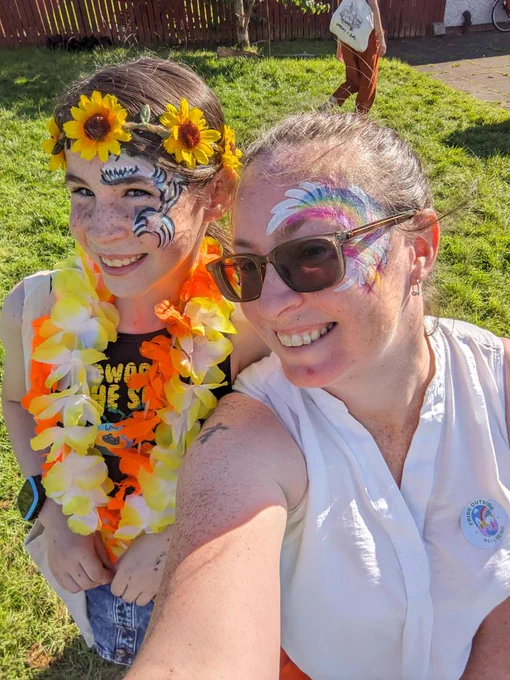 Ivy and I are smiling for a selfie on a sunny day. We both have painted faces - Ivy has a shark and I have a sort of rainbow explosion. Ivy also has a sunflower crown and two floral garlands around her neck. 