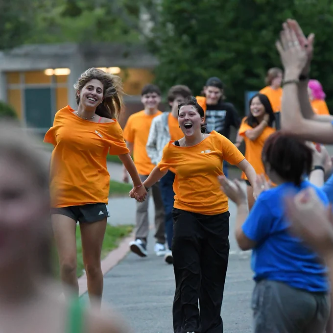Two students, wearing yellow shirts hold hands and skip down a line of clapping onlookers. 