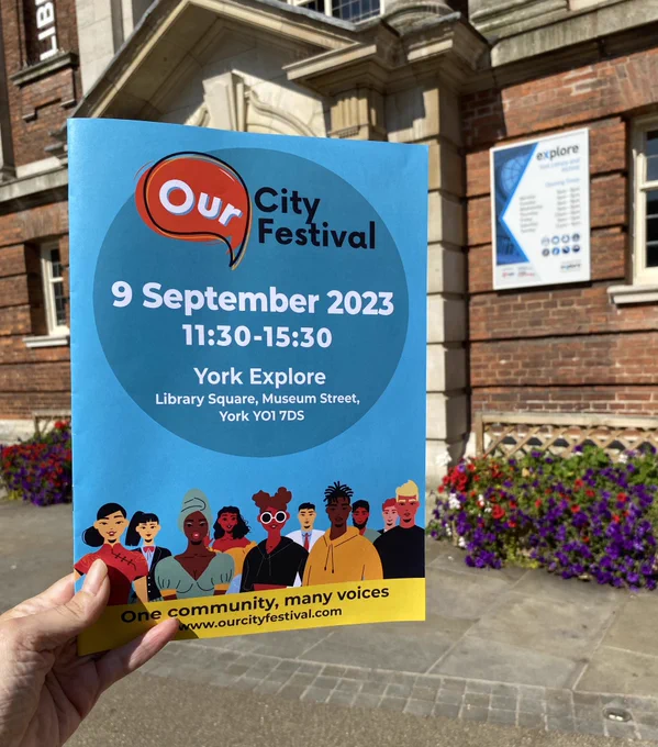 A leaflet that is predominantly blue with the words ‚Our City Festival, 9 September 2023, 11:30-15:30, York Explore, Library Square, Museum Street, York’ on the front. The leaflet is being held up in front of the entrance to York Explore Library.
