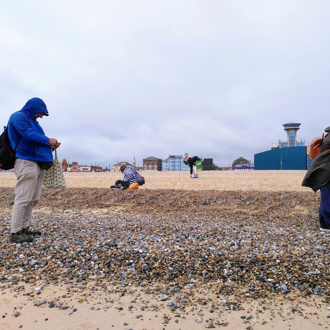 a group of adults are exploring a bank of pebbles on Great Yarmouth beach. They are standing, looking and crouching down. In the background, the amusements are buildings can be seen and it's a cloudy day