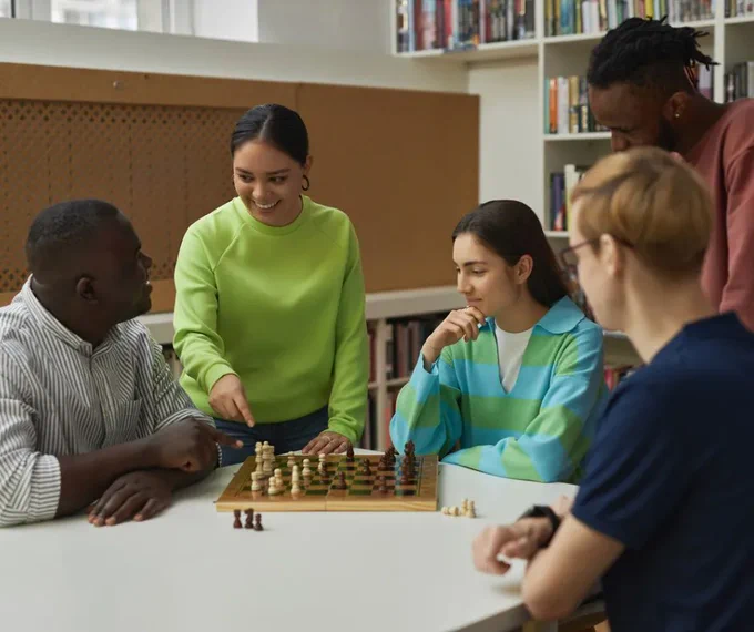 Group of five young people hanging out around a chess board mid-game. Two people are pointing at pieces and seem like they're engaged in conversation while the other three ponder the board state.