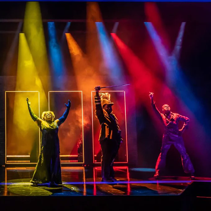 Six people stand on a stage in a line in extravagant poses. Behind them are the outline of six doors with neon tube lighting outline. The lighting is harsh spots in rainbow colours, starting with red on the right and ending with purple on the left. The doorways are lit those colours too. The rest of the stage is fairly dark.
