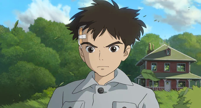 A film still from The Boy and the Heron. A boy with a bandage on his head aggressively stares forward as the wind blows leaves behind him. There is also a house in a field with trees behind him. 