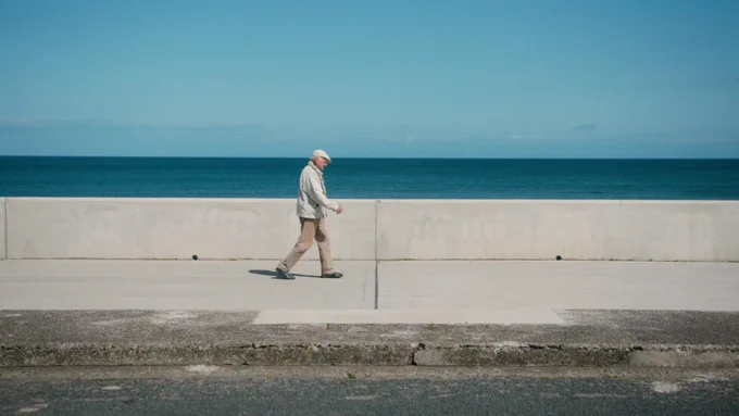 a film still from Grandad and The Pier featuring an older man dressed in white and cream walking easterly along a concrete sea defence with a blue sky and sea beyond | image: Reuben   Jamie 