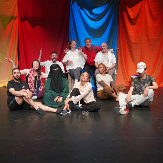 A group of people (making Guest House - Creating Together / Displace Yourself) sat together, smiling in front of a colourful background of draped fabric. 