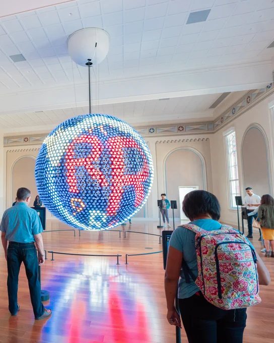 The Spoken World gallery at Planet Word. Museum visitors stand in front of tablets that are placed around the room, and the globe is activated with two large red letter Rs.