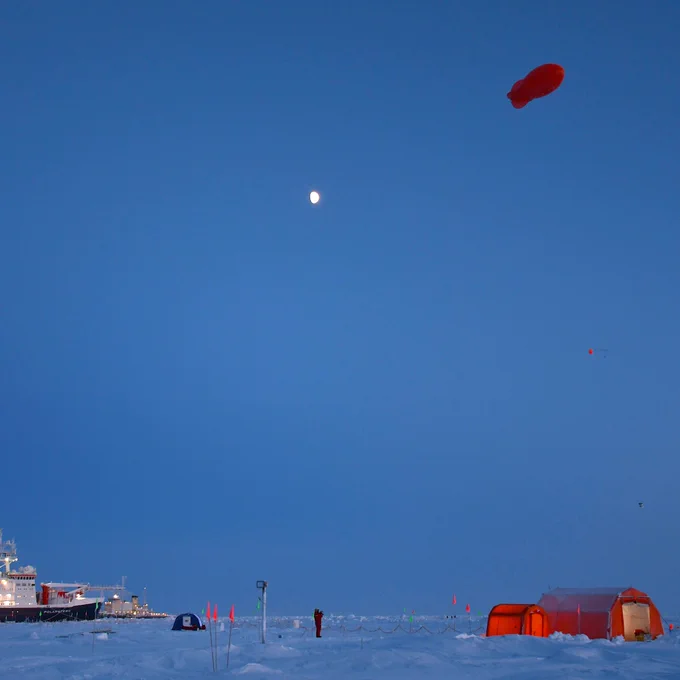 Scientists studying clouds on the RV Polarstern fly a weather balloon above the tents pitched on the ice. The balloon is like a red zeppelin. The sky is almost dark, and the orange tents glow with internal warm light. The RV Polarstern is visible in the distance.
