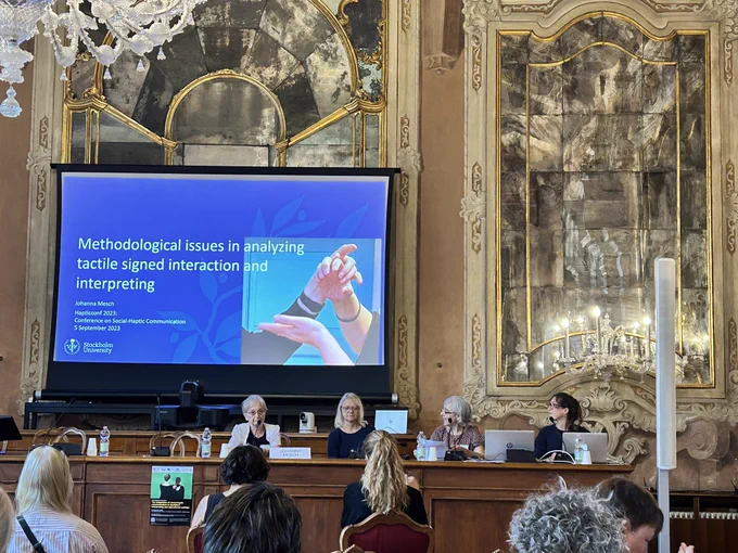 Photo of 4 people sat in front of a screen, one of whom is the presenter Prof. Johanna Mesch. The slide of the screen has an image of the hands of two tactile sign language users and the title of the presentation: Methodological issues in analysing tactile signed interaction and interpreting.