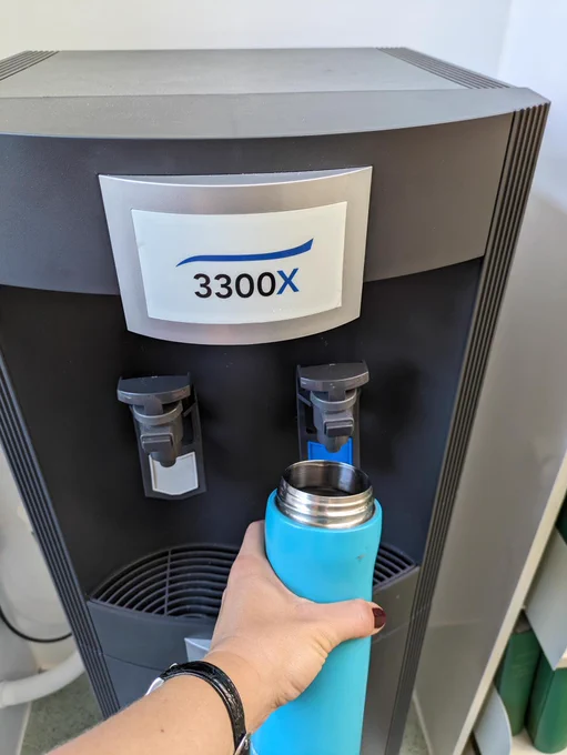 An image of a turquoise water bottle being held up to one of the two taps on the water cooler.