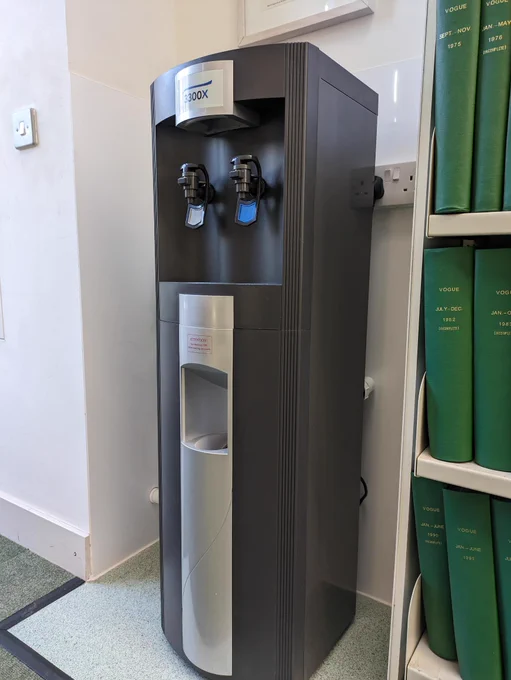 Image of a tall grey water cooler next to a bookcase of green leather bound volumes.