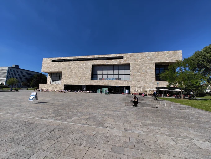Blue sky above the large stone brick building where the Euromat2023 conference is being held. A large stone pavilion leads up to the building with trees and grass on either side.
