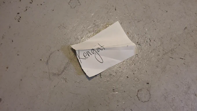 Betonilattialla paperilappu, jossa pelkästään sana "kengät".