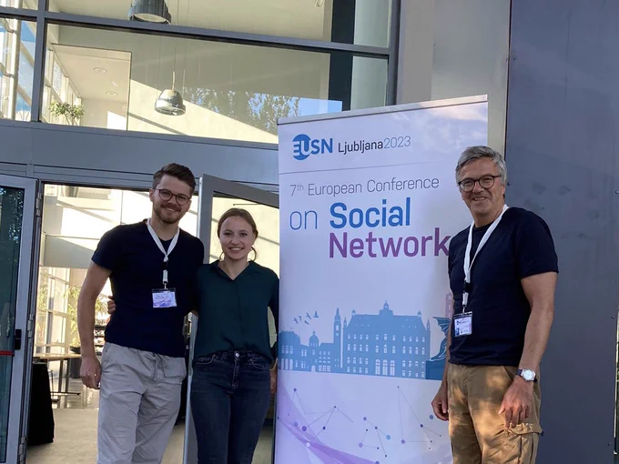 Cornelius Holler, Annabell Schüßler und Henning Plessner (vlnr) stehen vor einem Banner der 7. Europäischen Tagung über soziale Netzweke [7th European Conference on Social Networks]