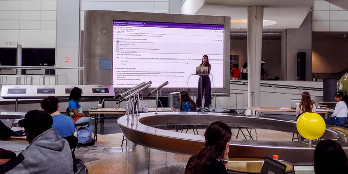 Safinah Ali stands at a podium in a lecture hall, with a large screen behind her showing a presentation. She is teaching machine learning and data science concepts to high schoolers in the SRMP program. Credit: AMNH