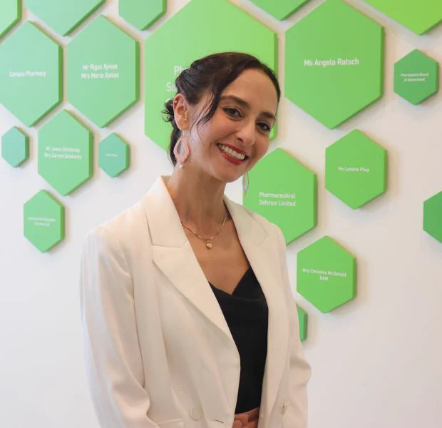 A woman in a black top, white jacket & pink earrings stands smiling side-on in front of a white wall with multiple green hexagon signs.