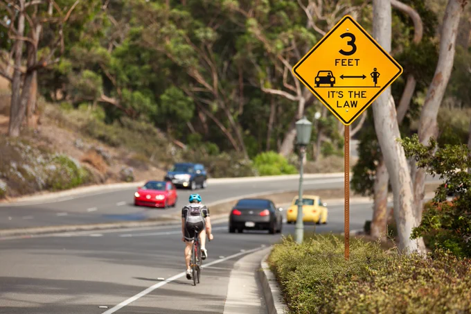 Bicyclist riding on a road. Sign next to the road reads "3 feet <clip art style drawing of a car and a cyclist with a double-sided arrow in between> It's the law."